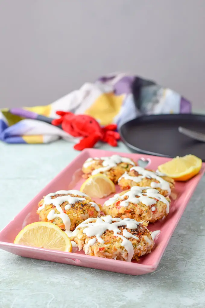 Crab cakes on a pink platter with sauce drizzle on top and lemon wedges around them!