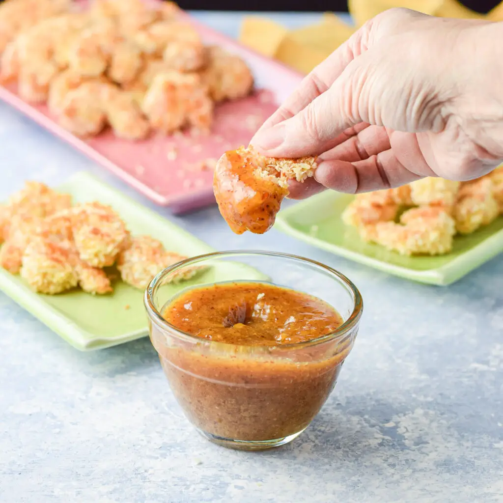 A hand holding a baked shrimp that has been dipped in apricot sauce. There are plates of coconut shrimp in the background