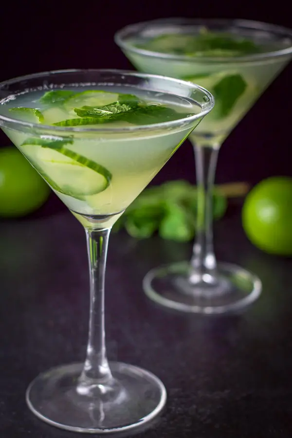 Two cucumber martinis poured out with some limes and mint in the background