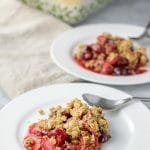 two plates of cherry rhubarb crisp with the baking dish of crisp in the background