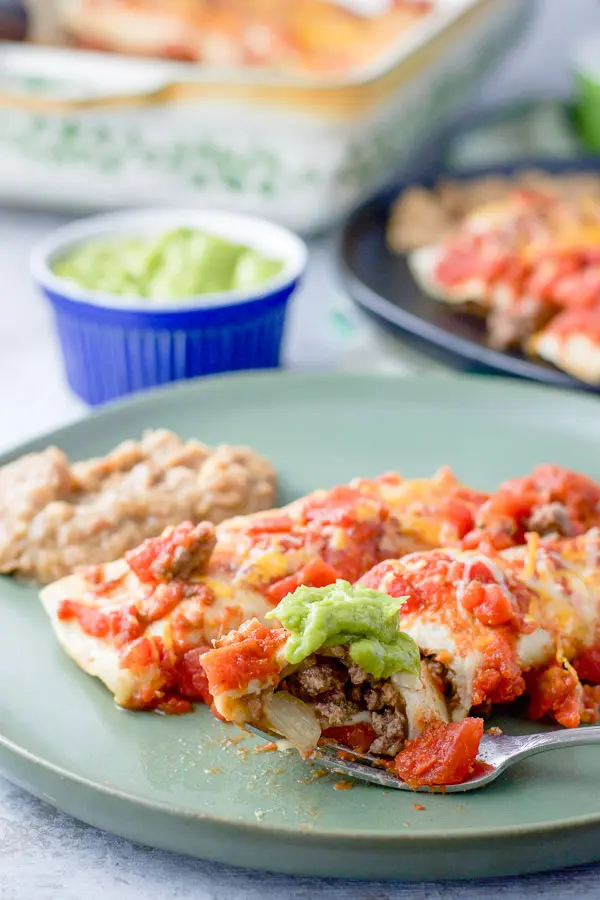 Forkful of enchilada on the plate with two enchiladas and a serving of refried beans. In the background is guacamole and another plate of enchiladas