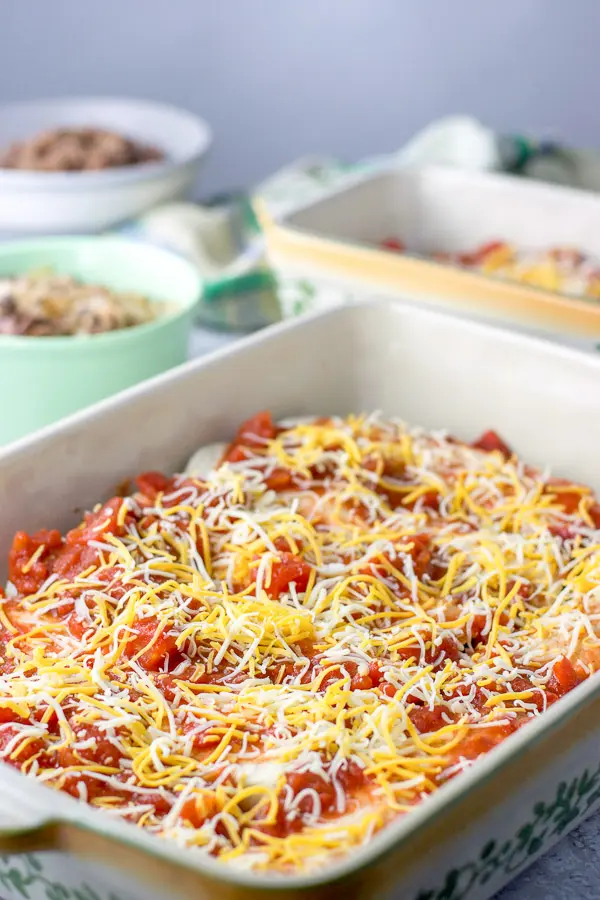 Cheese sprinkled on the enchiladas that are in a baking dish with another baking dish and beef in the background
