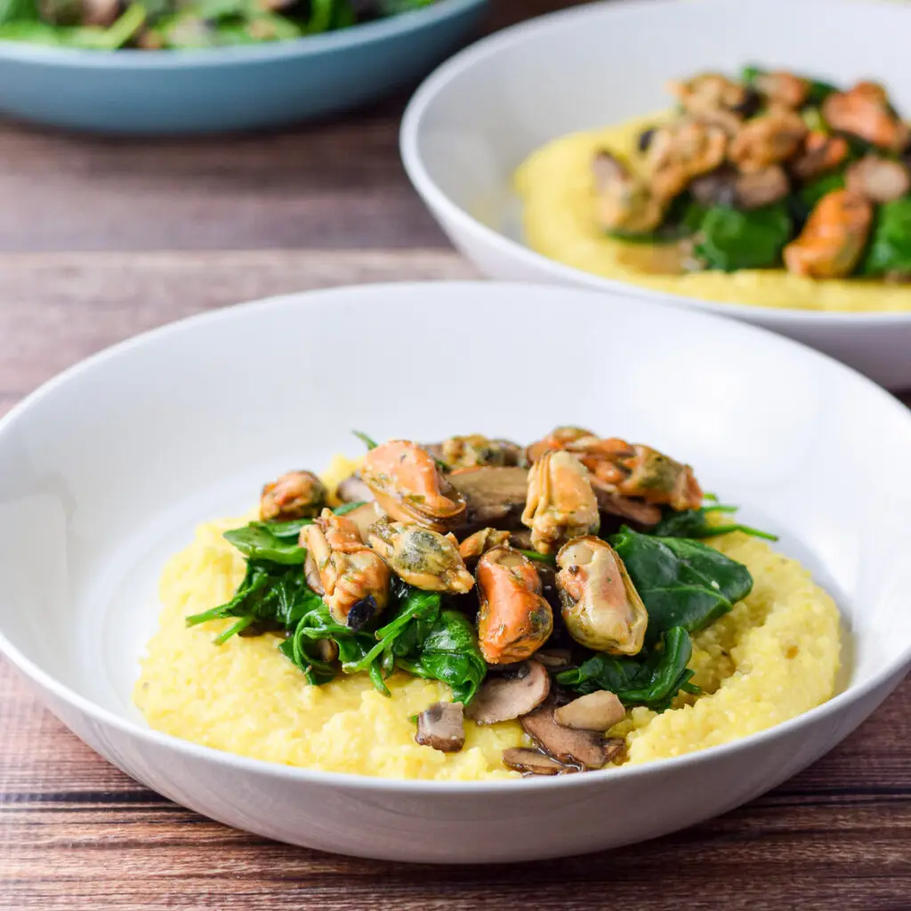 Two shallow white plates with mussels, spinach and mushrooms on polenta - square