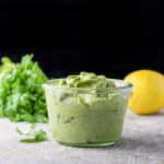 A jar filled with a green dressing with cilantro and lemon in the background