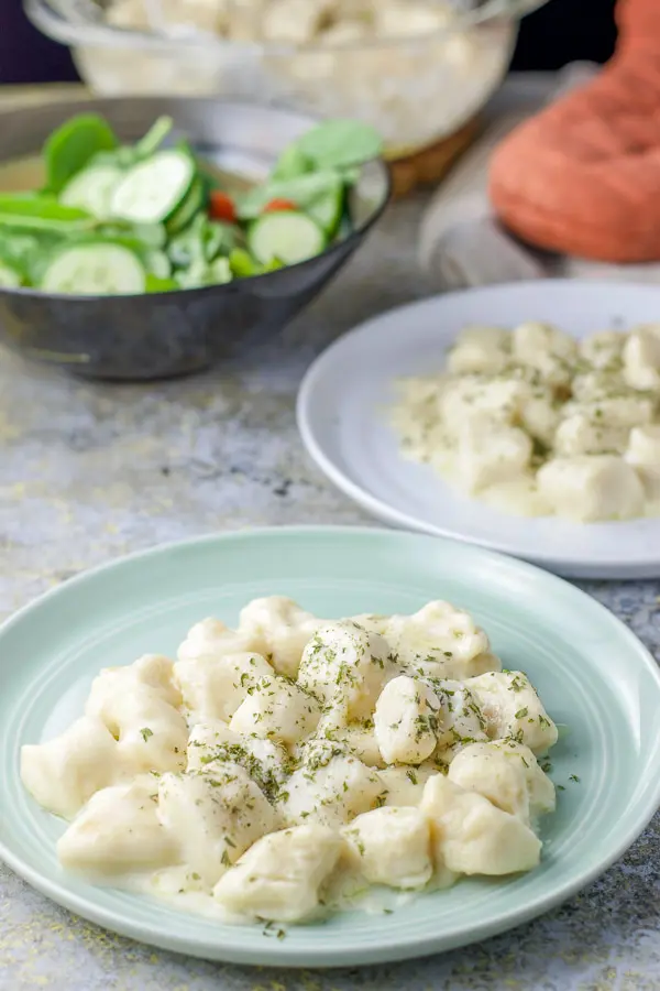 A green round plate with gnocchi with cream sauce on it with another plate, salad and casserole dish in the background
