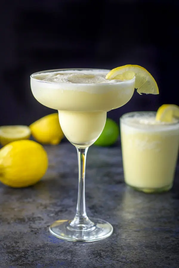 Classic tall margarita glass filled with lemon margarita, short glass with margarita in back, surrounded by lemons and a lime