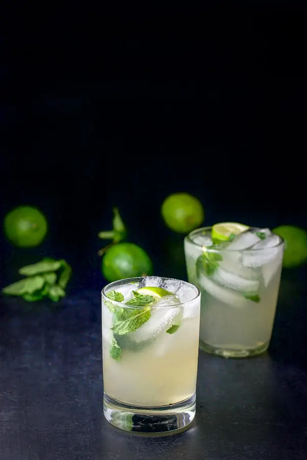 Two double old fashioned glasses filled with the mojito with a bunch of limes and mint leaves in the background