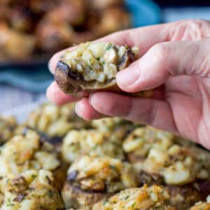 A female hand holding a stuffed mushroom with a bite taken out. There is a big plate of mushrooms underneath