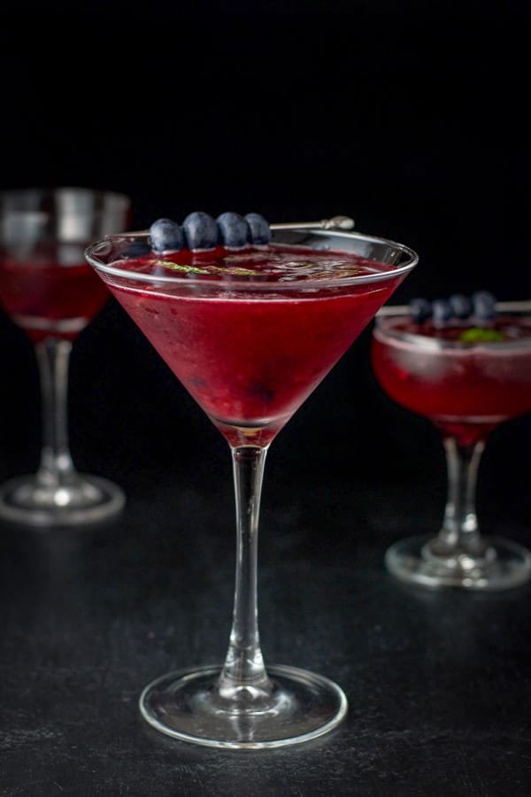 Blueberry cosmo with blueberries and mint leaves for garnish in martini glass in front and a coupe glass in back
