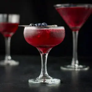 Three glasses of the blueberry cosmo in vertical view withe the coupe glass in front - square