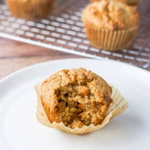 Close up of a white plate with one of the muffins with a bite taken out of it. There are more muffins in the background on a rack - square