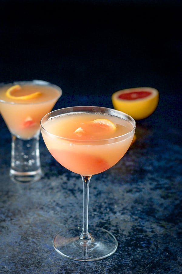 Bowl shaped glass filled with the cosmo in front of the bubble martini glass. There is also some cut grapefruit in the background