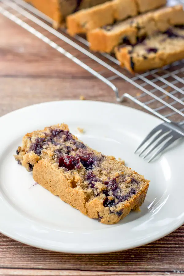 A piece of blueberry bread on a plate with a few pieces on a wire rack behind it