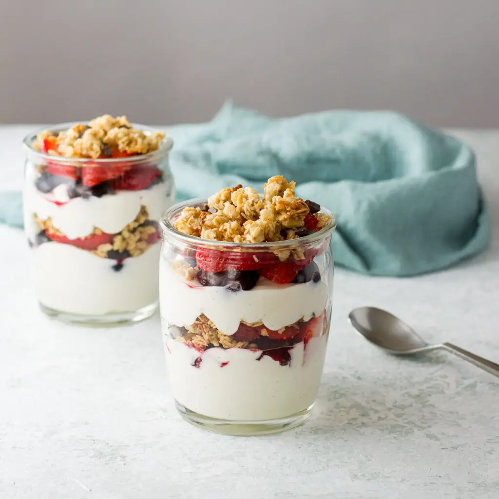 Two jars filled with layered yogurt, fruit and granola with a napkin in the background - square