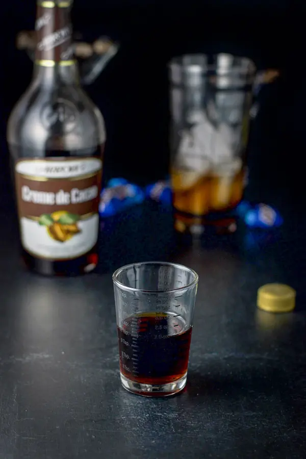 Creme de cacao poured out with the bottle, shaker, candy bars and glassware in the background