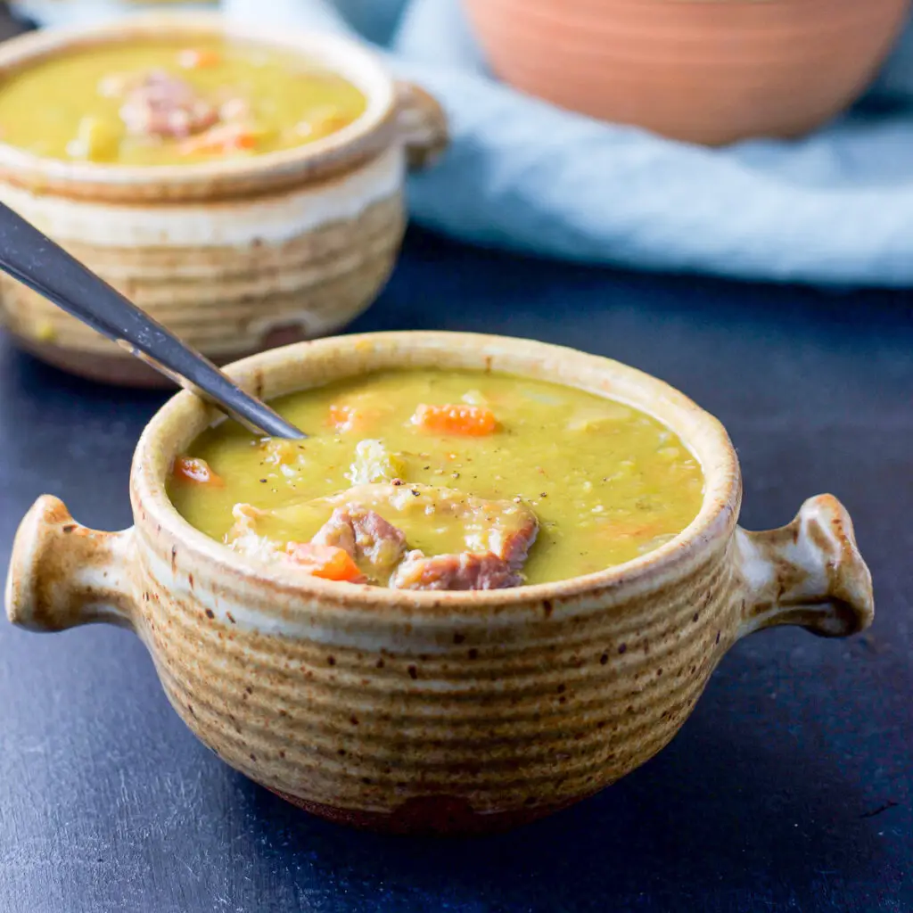 Two crocks filled with pea soup with a spoon in the front one. There is ham and carrots on the top of the soup - square