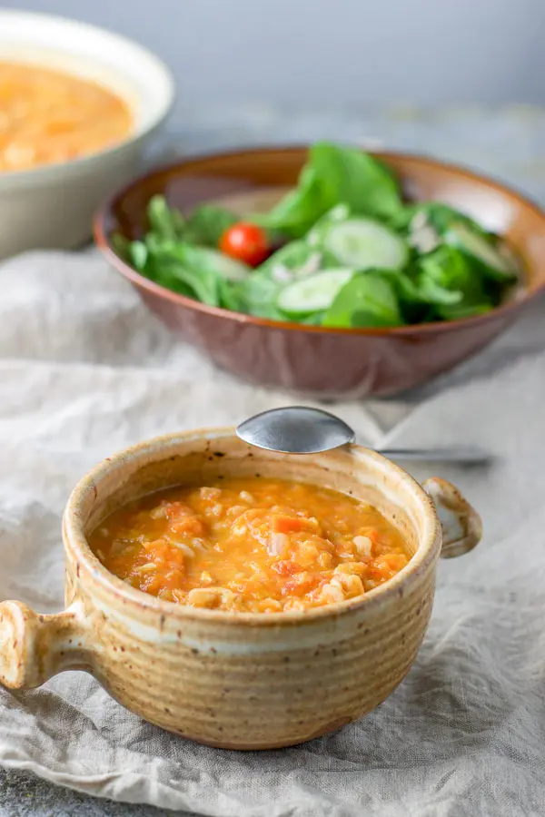White bean soup ladled into a crock with a spoon on the side of the bowl and the big bowl of soup and a salad in the background