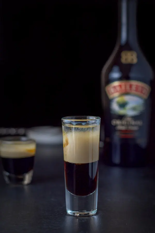 Baileys layered into the glass with the bottle in the background along with a white dish and spoon