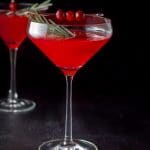 Cranberry cocktail in two martini glasses with the curved glass in front and garnished with cranberries and fresh rosemary