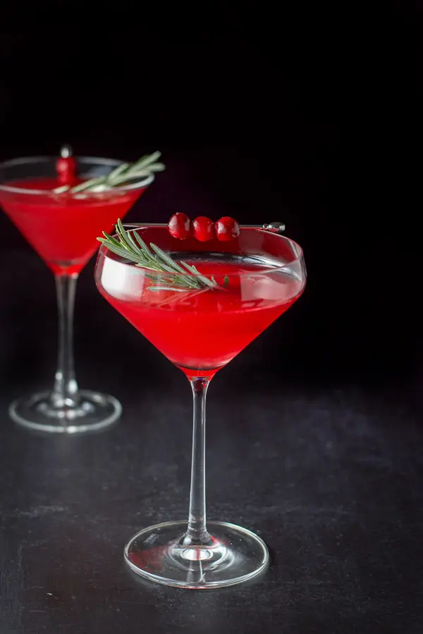 Fun curved martini glass filled with the red cosmo with fresh rosemary as a garnish along with some cranberries on a pick
