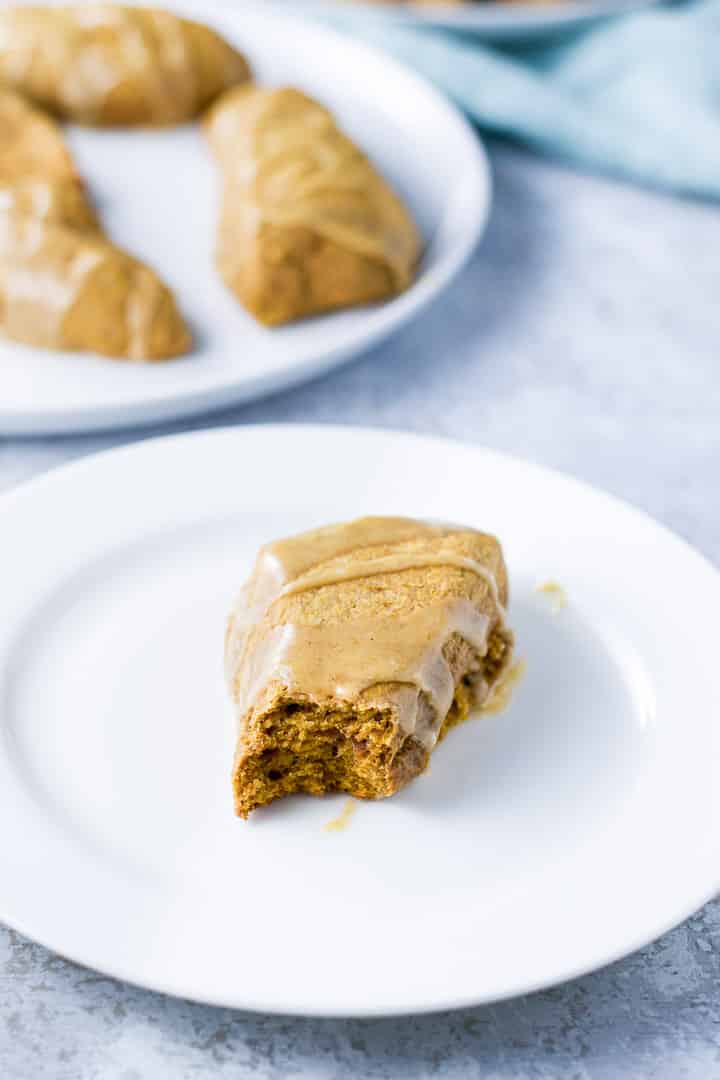 A white plate with a scone with a bite taken out of it and a plate of scones in the background