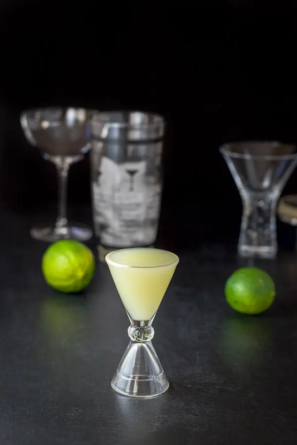 Lime juice poured out with a few limes, shaker and glasses behind it