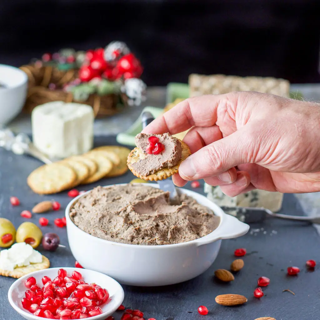 A male hand holding a cracker with liver pate on it with lots of crackers, cheese, pomegranate seeds and other appetizers - square