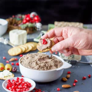 A male hand holding a cracker with liver pate on it with lots of crackers, cheese, pomegranate seeds and other appetizers - square