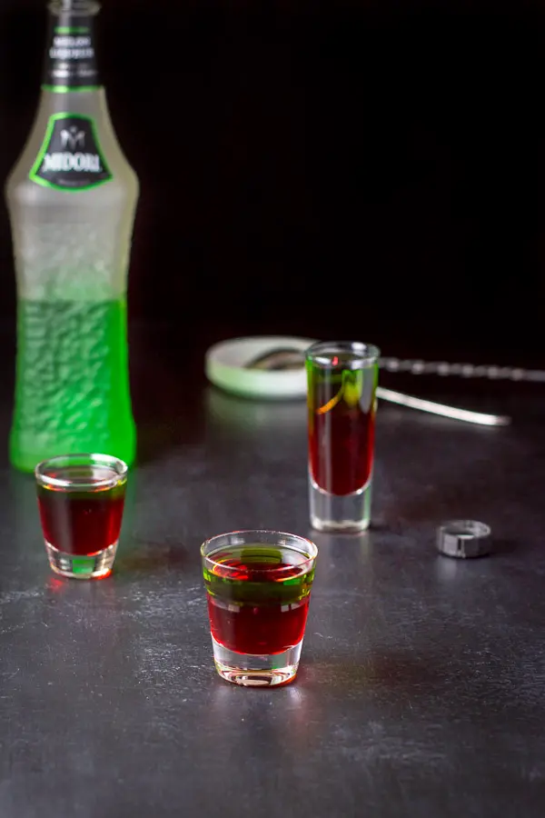 The Midori layered into the shot glasses with the bottle and spoons in the background