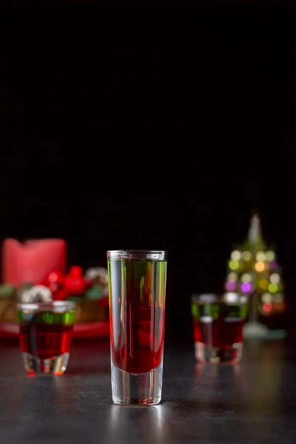 Vertical view of the tall glass filled with the green and red shot. There is a Christmas tree and candle in the background