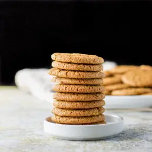 A tower of molasses cookies on a small white plate with a bigger plate filled with the cookies in the background