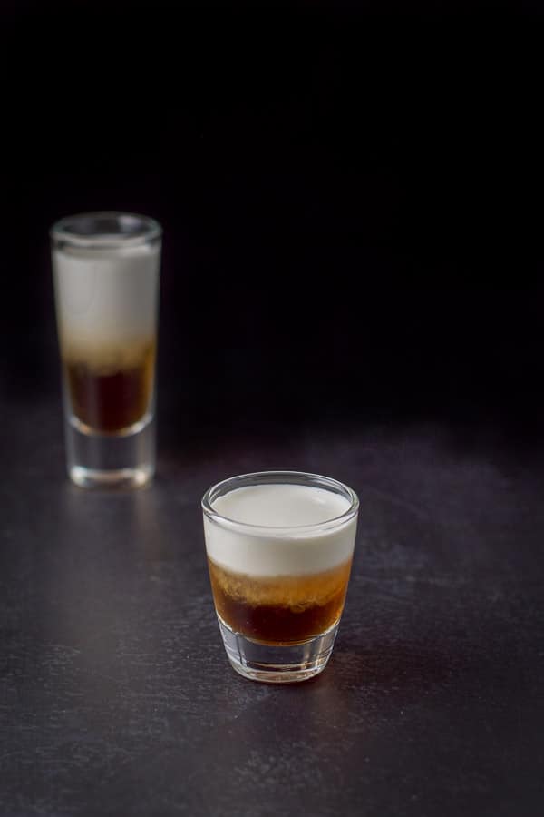 View of the short glass in front of the tall glass filled with the layered shot