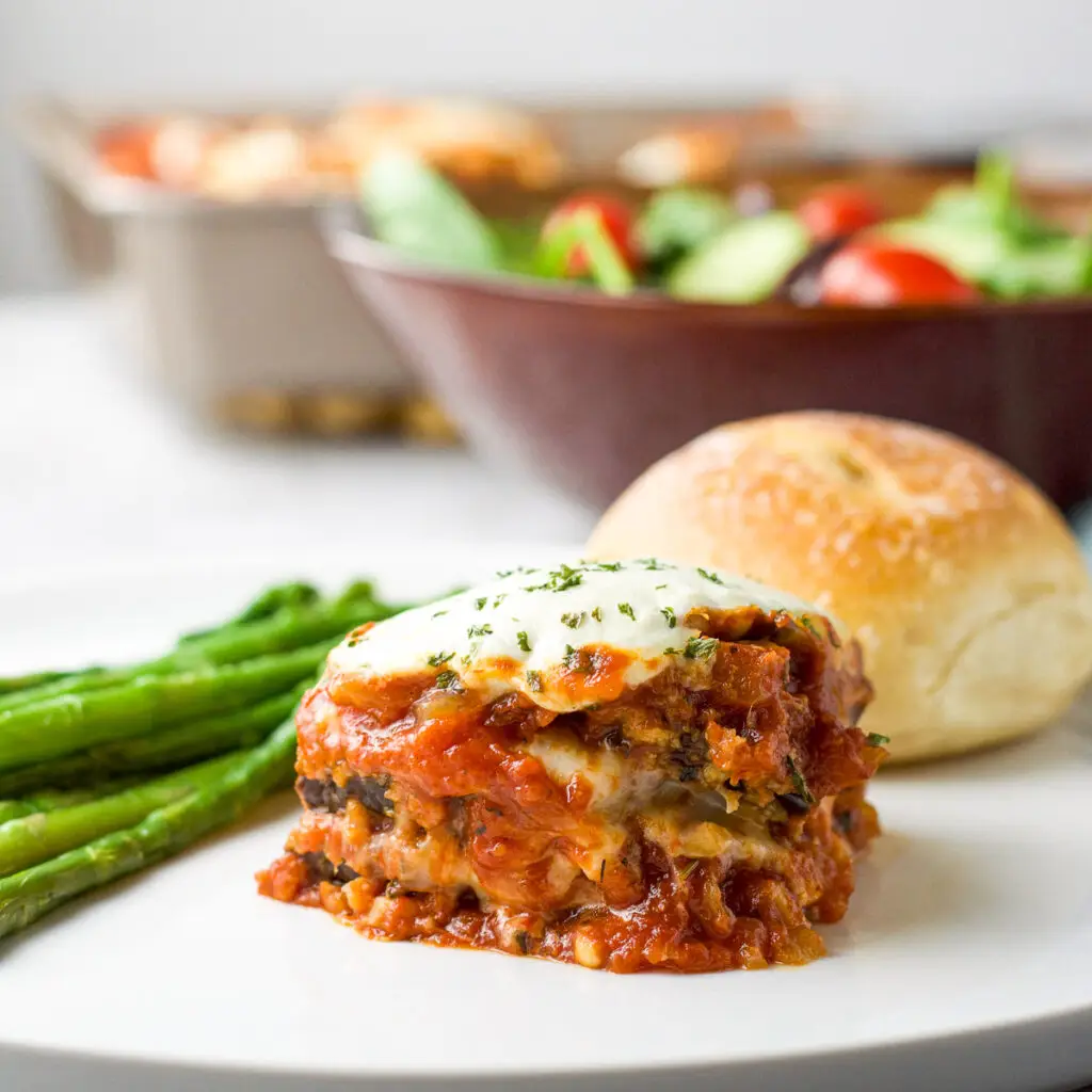 A piece of eggplant parmesan on a plate with asparagus and a roll - square