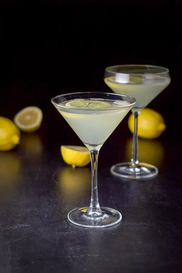 A classic martini glass filled with the lemon drink with the curved martini glass and lemons in the background