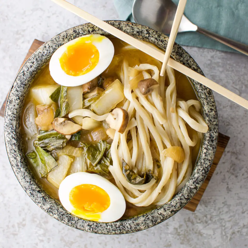 Square photo of the overhead view of a stone bowl with the noodle soup with an egg in it