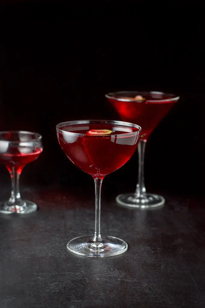 Lower photo of the red cocktail poured into 3 glasses - a bowl glass, classic martini and coupe. There are limes floating in the glasses