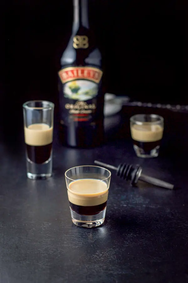 Baileys Irish cream layered on top of the Kahlua with the bottle in the background along with a pourer and cocktail spoon