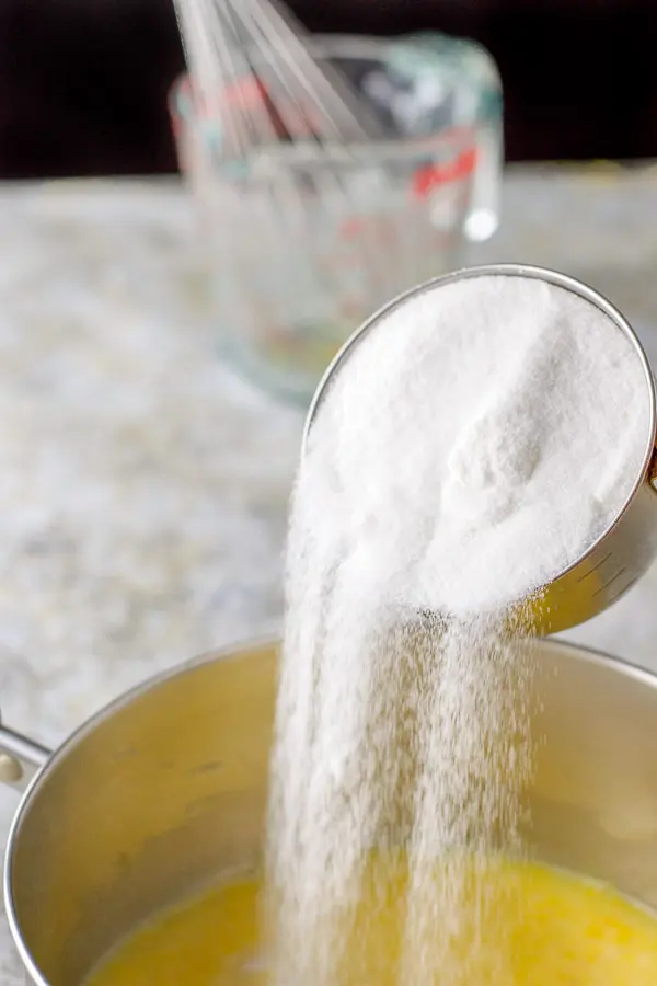 Sugar being poured into a pan of the lemon mixture with a glass measuring cup and whisk in the background
