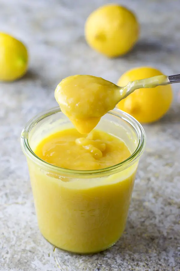 A dripping spoonful of curd over the jar with three lemons in the background