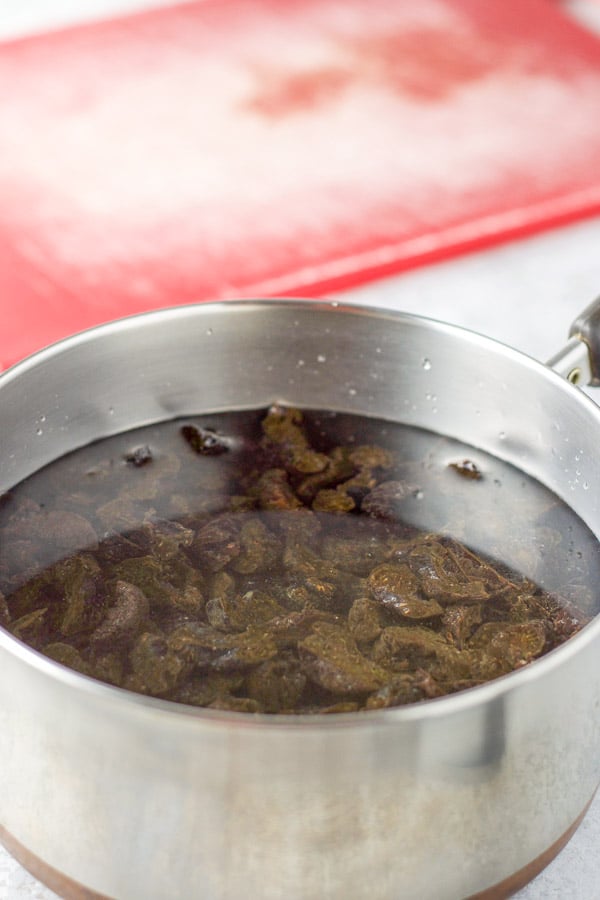 Water added to the prunes in a pan with the red cutting board in the background