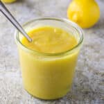 A spoon in a jar filled with lemon curd with a few lemons in the background - square