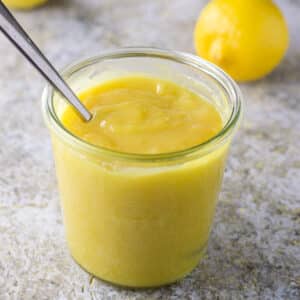 A spoon in a jar filled with lemon curd with a few lemons in the background - square