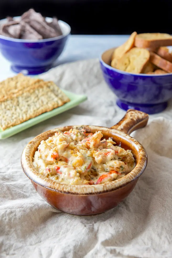 A brown crock filled with a dip with lobster poking out. There are crackers, chips and bread crisps in the background