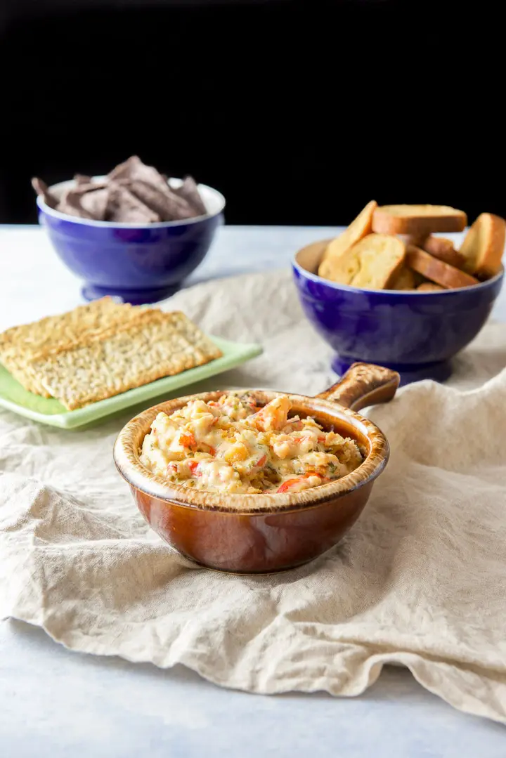 A brown crock filled with a chunky lobster dip with crackers, bread crisps and chips in the background