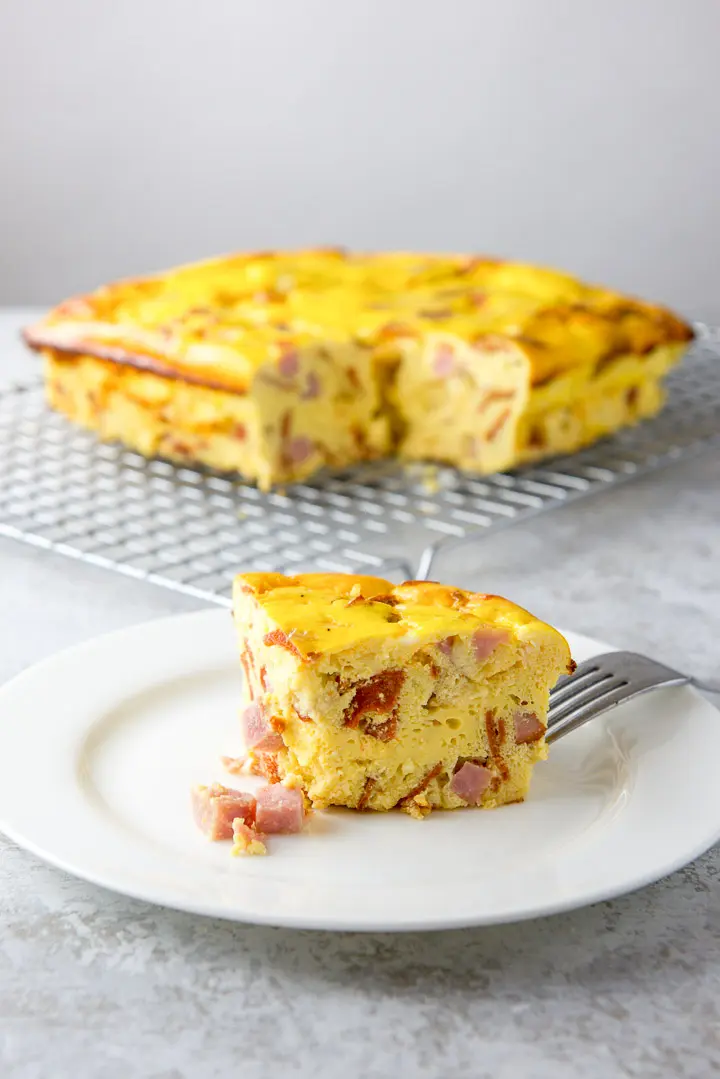 A slice of egg and ham loaf on a salad plate with the whole casserole on a cooling rack behind it.