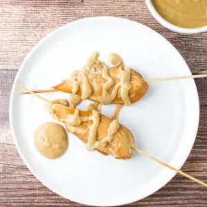 Overhead view of a white plate with two chicken skewers on it with peanut sauce dribbled on it - square