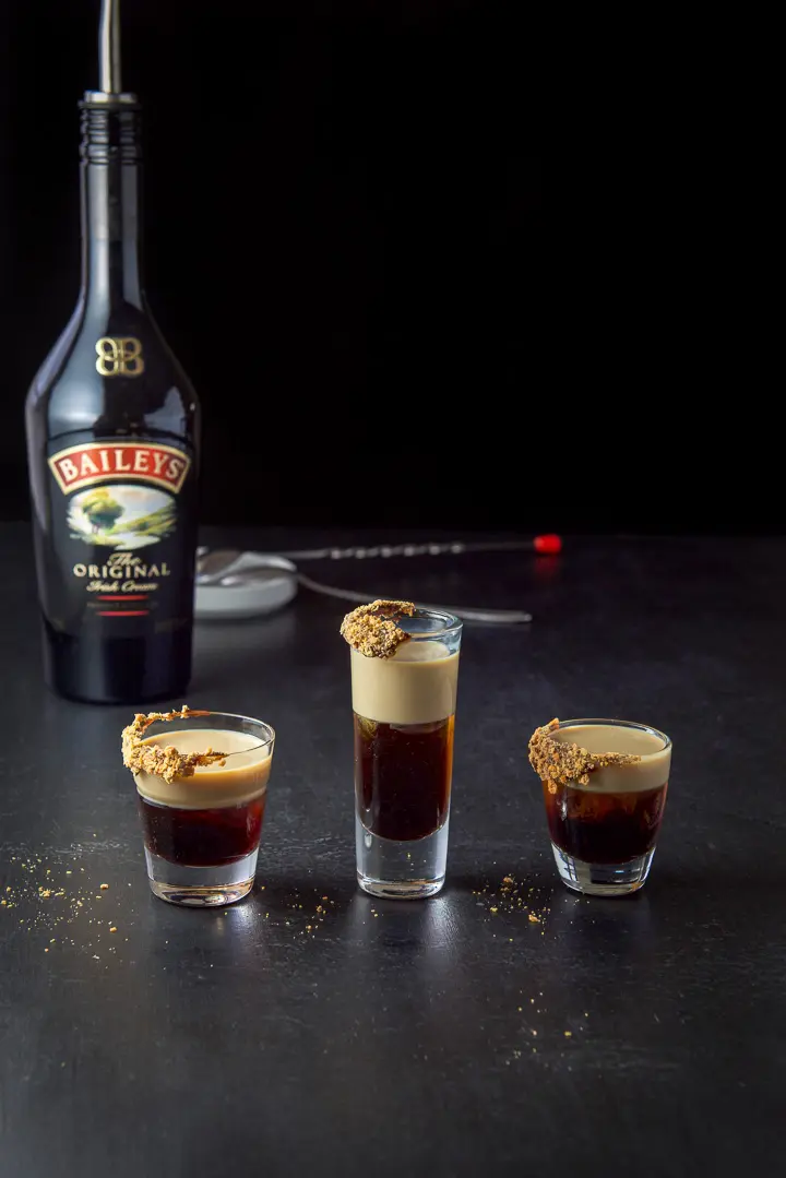Baileys layered in the candy bar garnished layered shot with the bottle, spoons and dish in the background