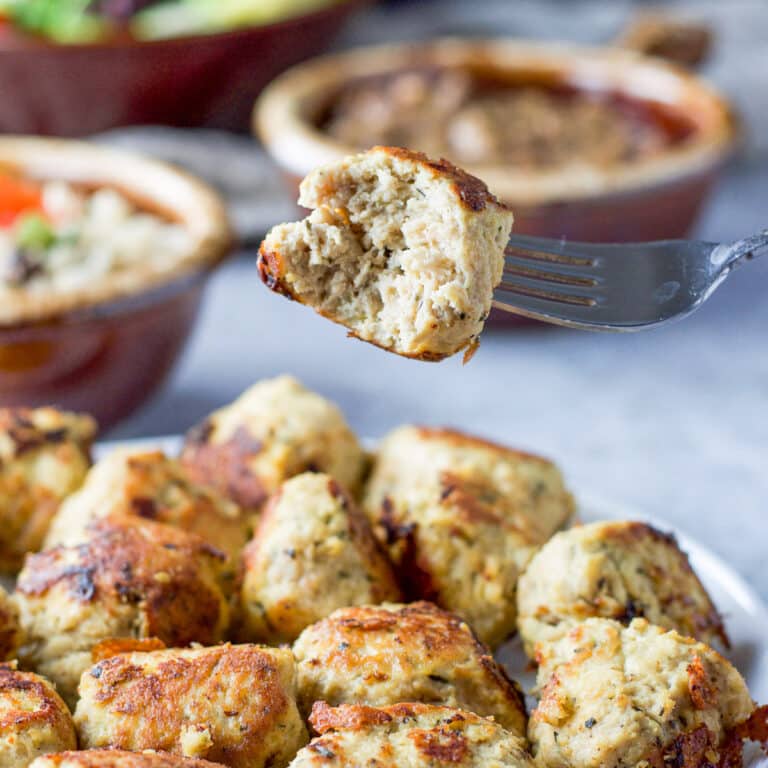 A fork with a meatball with a bite out of it held over a plate of meatballs. In the background there are beans rice and a salad in the background - square