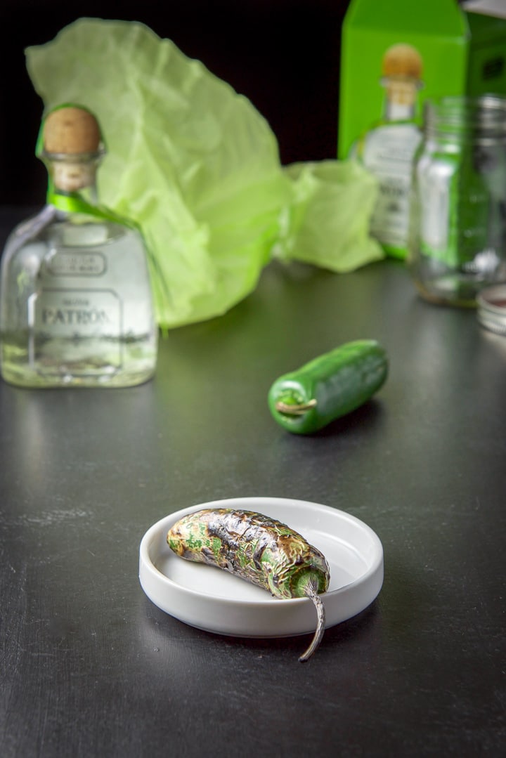 A roasted jalapeño in front and tequila and the box in the background