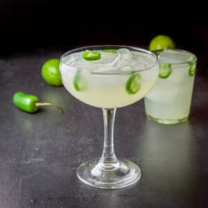 A bowl margarita glass in front of a rocks glass filled with the margarita with jalapenos in it and on the table